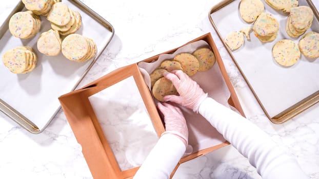 Flat lay. With precision, the woman is carefully arranging the sugar cookies, filled with dough-mixed sprinkles, into a rustic brown paper box.