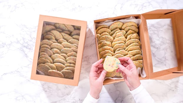 Flat lay. With precision, the woman is carefully arranging the sugar cookies, filled with dough-mixed sprinkles, into a rustic brown paper box.