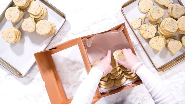 Flat lay. With precision, the woman is carefully arranging the sugar cookies, filled with dough-mixed sprinkles, into a rustic brown paper box.
