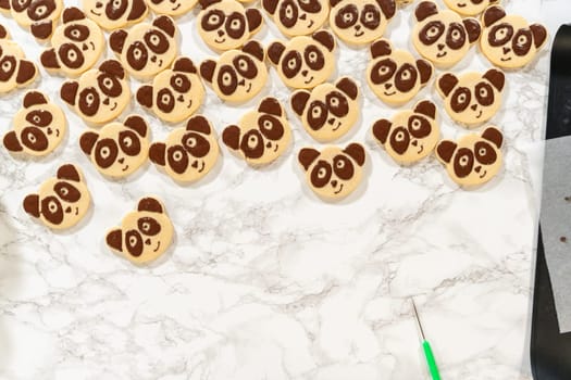Overhead view of undecorated and panda-face decorated shortbread cookies, with two piping bags filled with chocolate royal icing, arranged on a marble surface.