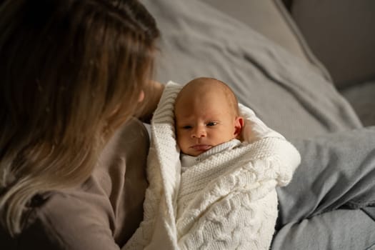 A mother lovingly gazes at her sleepy baby, who is swaddled in a soft white blanket. They enjoy a quiet afternoon in their warm living room.