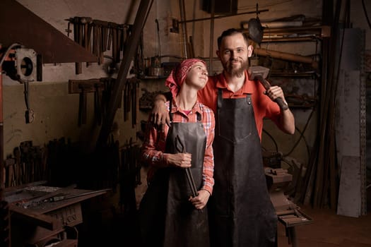 Professional blacksmiths in smithy portrait. Couple of young smiling european woman and bearded man hold hammer and metal tongs. Handmade metal work idea. Concept of handicraft and romantic love