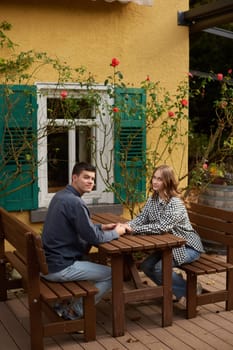 A serene garden scene featuring two individuals holding hands while sitting at a wooden picnic table. The picturesque setting includes a yellow building with green shutters, white-framed windows, and vibrant red rose bushes. The wooden deck and a barrel in the background add to the rustic charm of the outdoor patio area. The warm sunlight and blooming flowers create a peaceful atmosphere, perfect for a moment of connection. Two people holding hands at a garden picnic table outside a yellow building with green shutters, surrounded by roses