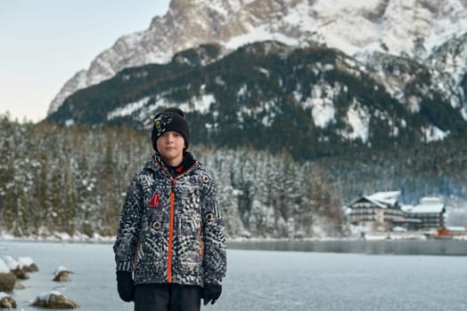 A young boy dressed in warm winter clothing stands on a frozen lake, surrounded by a breathtaking snow-covered landscape. Positioned slightly off-center to the left, he faces right, gazing into the distance. He wears a black beanie hat with a subtle logo, a stylish black and white patterned winter jacket with a red emblem on the chest, and black snow pants. His hands are clasped together in front of him, and he wears black gloves and snow boots, perfectly suited for the cold conditions. The background features a dense evergreen forest dusted with fresh snow, creating a stunning contrast of deep green and bright white. Towering snow-capped mountains rise in the distance, their peaks shrouded in light clouds, enhancing the depth and serenity of the scene. The frozen lake reflects the crisp blue sky, with scattered rocks and patches of open water adding texture. This peaceful and atmospheric winter setting captures the essence of solitude, adventure, and the beauty of nature, making it ideal for themes like seasonal travel, childhood exploration, outdoor adventure, and winter serenity. Young boy standing on a frozen lake in a snowy winter landscape with evergreen forest and majestic mountains in the background on a clear cold day