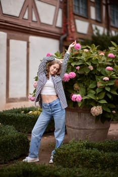 Capture the essence of autumn fashion and German countryside charm with this captivating outdoor portrait. A young woman, dressed in a trendy yet casual fall outfit, poses gracefully among vibrant blooming hydrangeas. The backdrop features a stunning traditional half-timbered house, exemplifying classic German architecture. The warm, rich colors of autumn create a cozy atmosphere, perfectly complementing the model's stylish ensemble. This image is ideal for fashion blogs, travel magazines, lifestyle publications, and autumn-themed marketing campaigns, showcasing the perfect blend of contemporary style and timeless rural beauty. A charming outdoor portrait of a young woman with wavy light brown hair, wearing a white cropped top, plaid flannel shirt, and blue jeans, posing confidently in front of vibrant pink and green hydrangea flowers. The background features a picturesque German half-timbered house, adding a touch of European architectural charm. The soft natural lighting and blurred background create a warm and inviting autumn atmosphere, making this image ideal for lifestyle, fashion, and seasonal themes. Perfect for use in blogs, magazines, travel guides, and advertising campaigns related to autumn fashion, cozy outdoor scenes, and European destinations. Young woman in casual autumn outfit posing among blooming hydrangeas in front of traditional half-timbered house in Germany – cozy and stylish outdoor portrait with warm fall colors