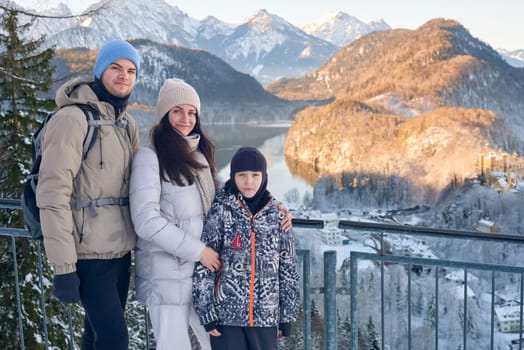 Adventurous Family Enjoying Mountain Sunset Panorama at Scenic Winter Lake Overlook with Snow Covered Peaks and Golden Alpine Glow - Heartwarming outdoor lifestyle portrait featuring happy active family of three enjoying breathtaking winter mountain landscape during golden hour sunset. Athletic parents and child wearing seasonal winter outerwear stand together at panoramic viewpoint overlooking pristine alpine lake surrounded by snow covered peaks and evergreen forest. Natural candid moment captures authentic family connection, outdoor adventure lifestyle, and spectacular wilderness scenery bathed in warm evening sunlight. Perfect for travel, outdoor recreation, family lifestyle, winter tourism, and adventure sports marketing.