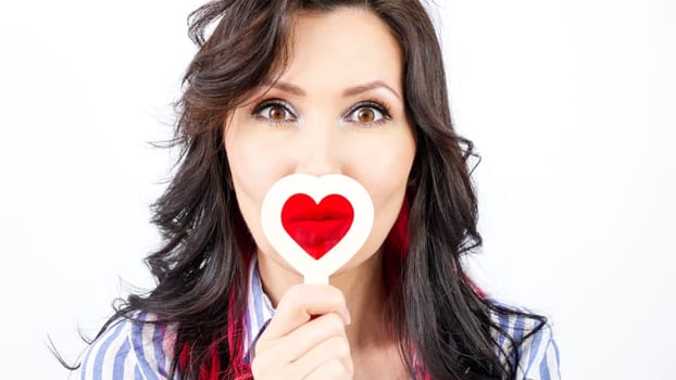 A young woman with long wavy brunette hair wears a blue and white striped shirt, showing a surprised expression while holding a heart-shaped sign in front of her mouth.