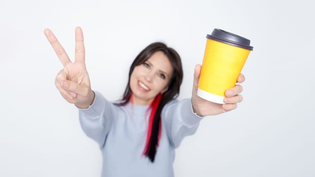 Smiling woman holding up a yellow mockup coffee cup with piece sign hand against a solid white background.