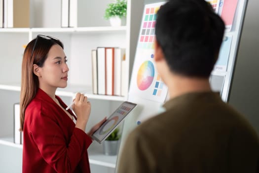 In a dynamic office setting, a female designer evaluates her color wheel on a digital tablet, engaging creatively with her colleague.