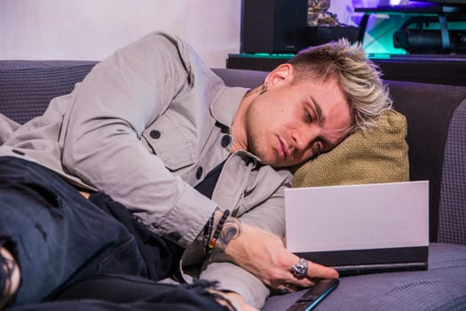 A young blond man with blue eyes is lounging on a couch, focused on reading a book. The relaxed atmosphere features warm lighting and comfortable decor, making it an inviting space.
