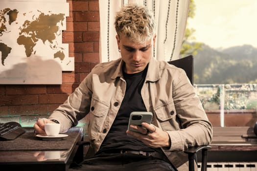 A young man with attractive blond hair and captivating blue eyes sits in a comfortable cafe, savoring a cup of coffee while focused on his smartphone. Natural light streams in through the window, enhancing the warm ambiance.