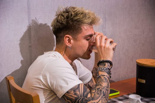 A young man with blond hair and blue eyes sits at a table, sipping a drink. Surrounded by a relaxed atmosphere, he showcases his tattoos and casual style while enjoying his moment.