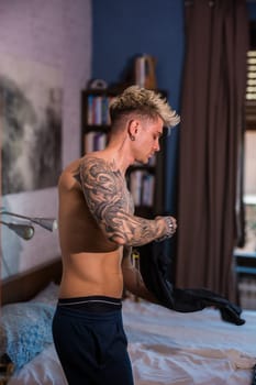 A young man is getting ready in a warmly lit bedroom. He is shirtless, showcasing his tattoos, as he puts on a black shirt while surrounded by comfortable decor.