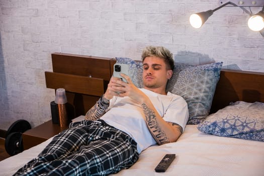A young man with blond hair and tattoos lies comfortably on his bed, absorbed in his smartphone. The cozy room features soft lighting and a casual vibe, perfect for relaxation.