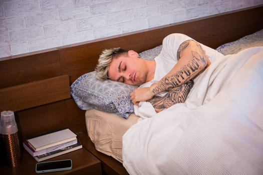 A young man with blond hair and blue eyes sleeps soundly in a neatly made bed. The soft morning light filters in, creating a tranquil ambiance for relaxation and comfort.