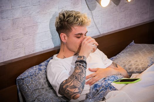 A young man with blond hair and blue eyes is seated on his bed, sipping from a glass. He has visible tattoos on his arms and appears relaxed in a cozy bedroom setting.