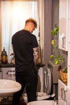 A handsome young man with blond hair and blue eyes stands in a bathroom, facing the toilet. Sunlight filters through the window, illuminating the space and creating a relaxing atmosphere.