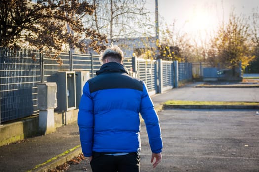 A young man with blond hair and blue eyes walks away in a blue and black jacket. The sun sets behind him, creating a warm atmosphere in a quiet urban area.