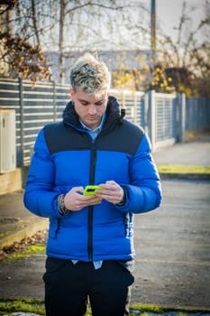 A handsome young man with blond hair and blue eyes stands outdoors, focused on his phone. He wears a vibrant blue jacket and appears to be in an urban environment during the day.