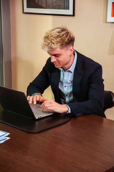 A handsome young man sits at a desk focused on his laptop. He has blond hair and blue eyes, dressed in a suit, clearly engaged in his work environment during daylight hours.