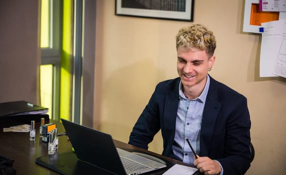 A young man with attractive features sits in a sleek office, focused on his laptop screen. He wears a navy blazer and smiles as he holds a pen, surrounded by papers and office supplies.