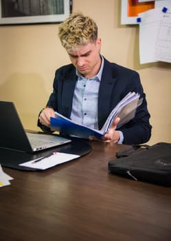 A young man with blond hair and blue eyes concentrates on reviewing a stack of documents at a modern office desk. The setting indicates a professional atmosphere, reflecting his diligent work ethic.