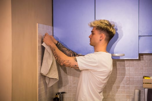 A young man with blond hair and blue eyes arranges a towel on the wall in a stylish kitchen. He is focused on the task while showcasing his tattoos and casual attire.