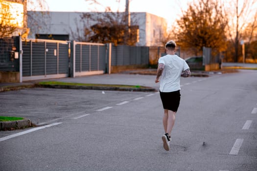 A young man with blond hair and blue eyes jogs down an empty street. The warm glow of sunset highlights the tranquil urban landscape with trees lining the path.