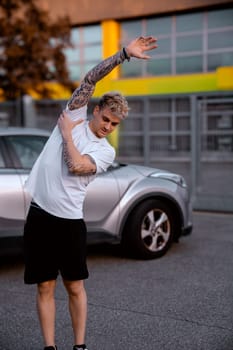 A handsome young man with blond hair and blue eyes stretches his arms while standing next to a silver car in an urban area during sunset. His athletic appearance highlights an active lifestyle.
