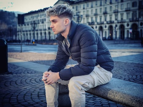 A young man with attractive features sits pensively on a stone bench in a quiet urban setting. His blond hair and blue eyes stand out against the backdrop of buildings during daylight.