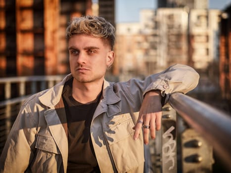 A young blond man with blue eyes leans against a railing in an urban setting at dusk. His stylish outfit and relaxed posture convey confidence as the city lights begin to glow.