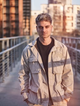 A handsome young man with blond hair and blue eyes stands on a contemporary urban bridge. The warm glow of sunset highlights his features, enhancing the stylish look of his casual outfit.