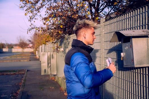 A young, attractive man with blond hair and blue eyes stands beside a mailbox. He holds letters and looks pleased to find his mail on a clear, sunny day in a peaceful area.