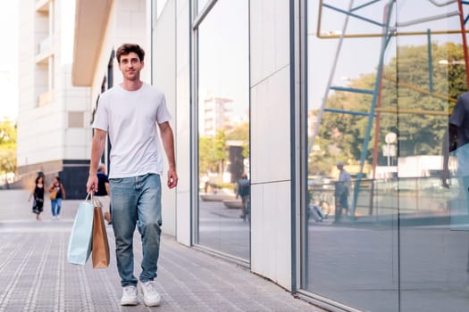 young adult man in casual clothes walking down a sidewalk near of a mall carrying shopping bags, concept of youth and urban lifestyle, copyspace for text