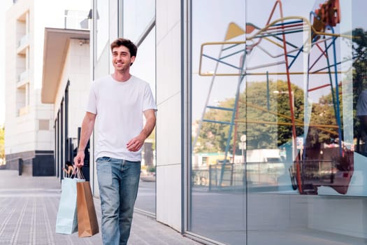 young adult man in casual clothes walking down a sidewalk near of a mall carrying shopping bags, concept of youth and urban lifestyle, copyspace for text