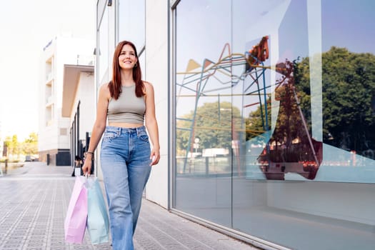 young adult woman in casual clothes walking down a sidewalk near of a mall carrying shopping bags, concept of youth and urban lifestyle, copyspace for text