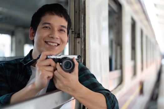 Excited about capturing memories, a young man smiles while holding his camera from a train window, showcasing his adventurous spirit.