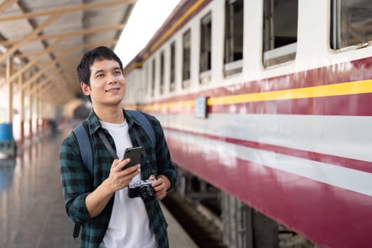 A cheerful young man at a train station, capturing the excitement of upcoming travels with a smartphone in hand and a camera ready to document his adventure.