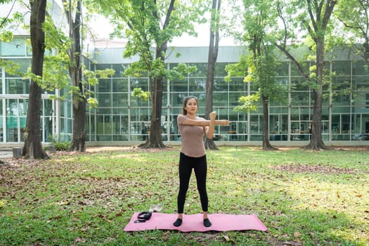 Prioritizing fitness, a woman stretches her arm while standing on a yoga mat, surrounded by equipment, in a vibrant outdoor setting.