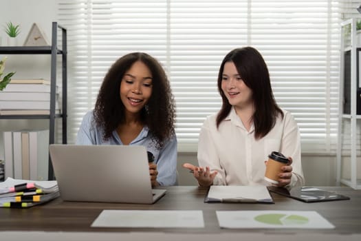 Two friends at work laugh and discuss ideas over coffee and a laptop, showcasing a positive and encouraging collaborative atmosphere.