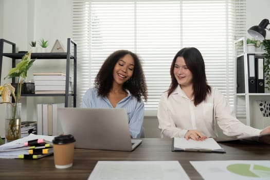 In a bright office, two women share insights while reviewing digital content on a laptop, fostering a productive and engaging work environment.