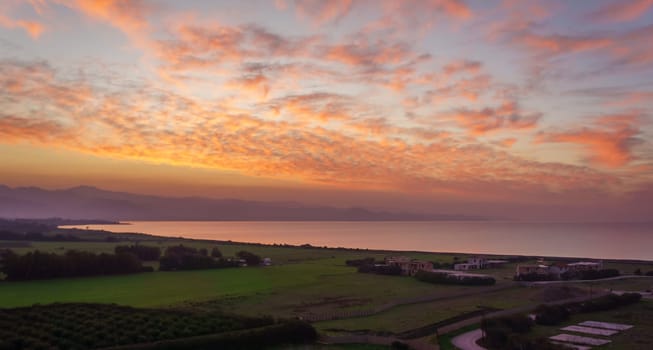 Vibrant hues fill the sky as the sun sets over calm waters and lush green fields. The peaceful atmosphere invites reflection and appreciation of nature's beauty.
