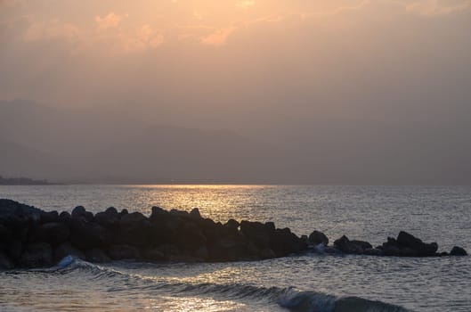 Golden hues blanket the horizon as the sun sets over tranquil waters. Waves gently lap against the rocky shore, creating a peaceful environment at dusk.