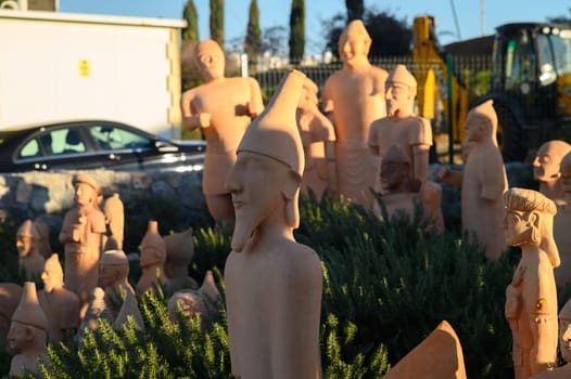 A diverse array of clay sculptures stands proudly in a sunlit outdoor space, showcasing various artistic styles and figures among greenery. The warm light highlights their intricate details.