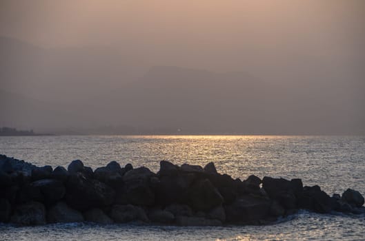 Golden hues shimmer across the calm waters as the sun sets behind the horizon, casting a serene glow over the rocky coastline and distant mountain silhouettes.
