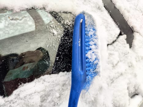 Using a brush to remove snow from a frozen car window on a cold winter morning in a snowy landscape.