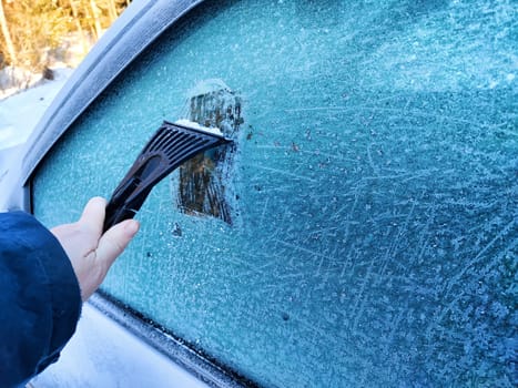 Person uses a scraper to remove frost from a car window on a chilly morning, surrounded by a winter landscape with snow.