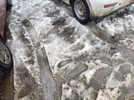 Melting snow covers the road in spring, showing wet patches and tire marks from cars driving through the slushy terrain.