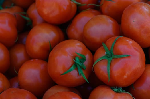 Vibrant red tomatoes are piled high, glistening in the sunlight at a bustling farm market. Their perfect round shapes and green stems create a colorful display of fresh produce.