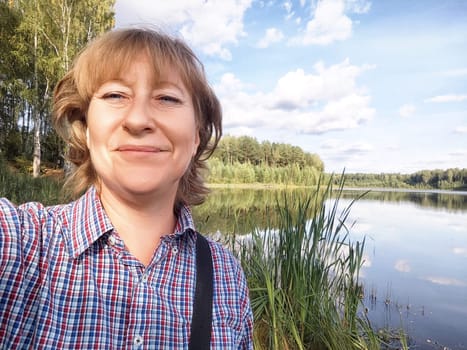 A middle-aged female traveler blogger takes a friendly selfie beside a tranquil lake surrounded by lush greenery.